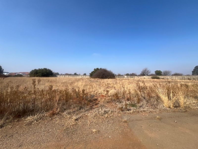 A vacant residential plot featuring dry grass, a perimeter wall, and an asphalt driveway entrance.