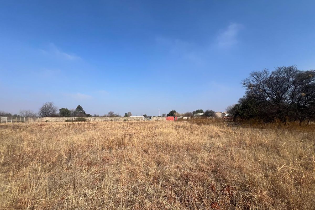 A wide-angle view of a vacant residential plot featuring dry grass and a perimeter boundary wall in the background.