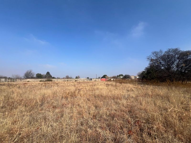 A wide-angle view of a vacant residential plot featuring dry grass and a perimeter boundary wall in the background.
