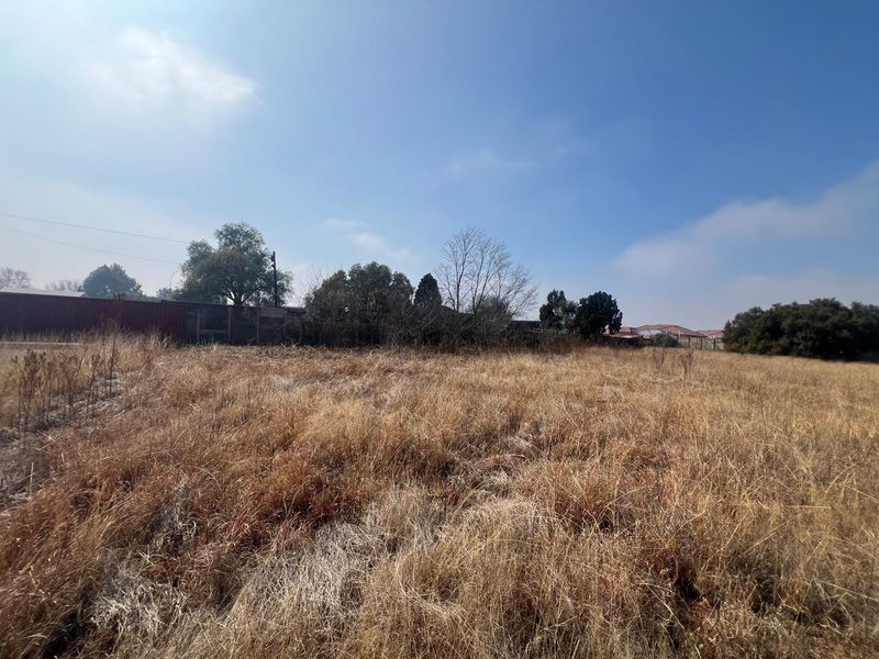 A view of the vacant residential land featuring dry grass, perimeter fencing, and mature trees in the background.