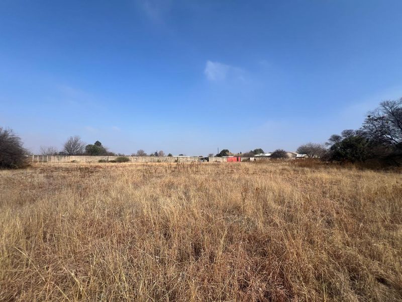 A wide-angle view of a vacant residential plot featuring dry grass and a perimeter boundary wall in the background.