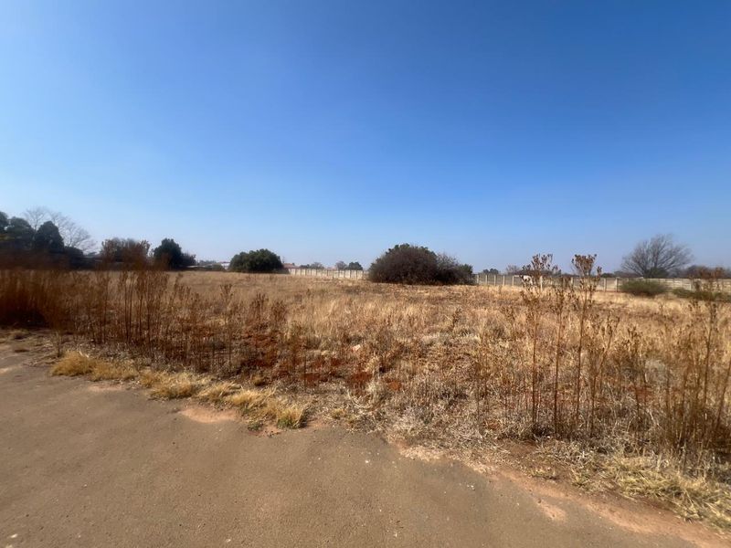 A vacant residential plot featuring dry vegetation, perimeter fencing, and direct access to a paved road.