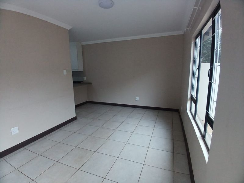 An empty open-plan living area featuring neutral tiled flooring, coved cornicing, and large aluminium-framed windows, with a glimpse of the adjacent kitchen space.