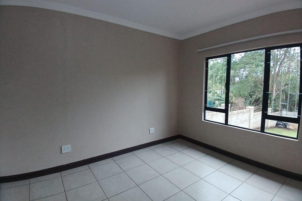 A clean, empty bedroom featuring neutral wall tones, tiled flooring, and large windows fitted with security burglar bars.
