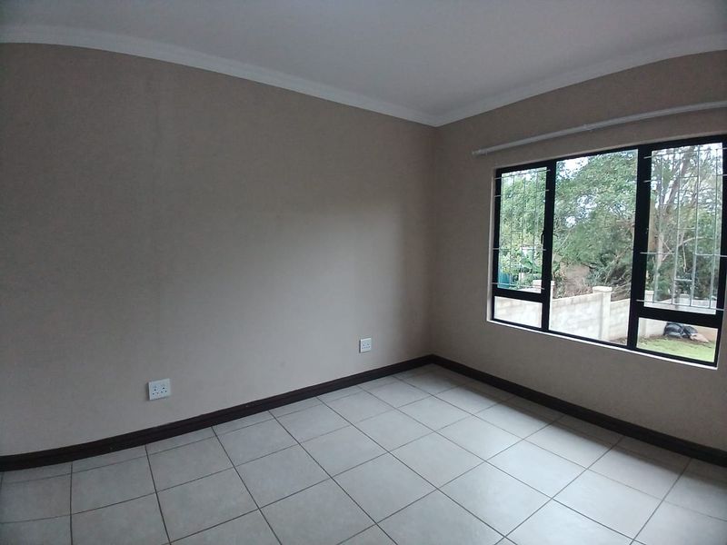 A clean, empty bedroom featuring neutral wall tones, tiled flooring, and large windows fitted with security burglar bars.