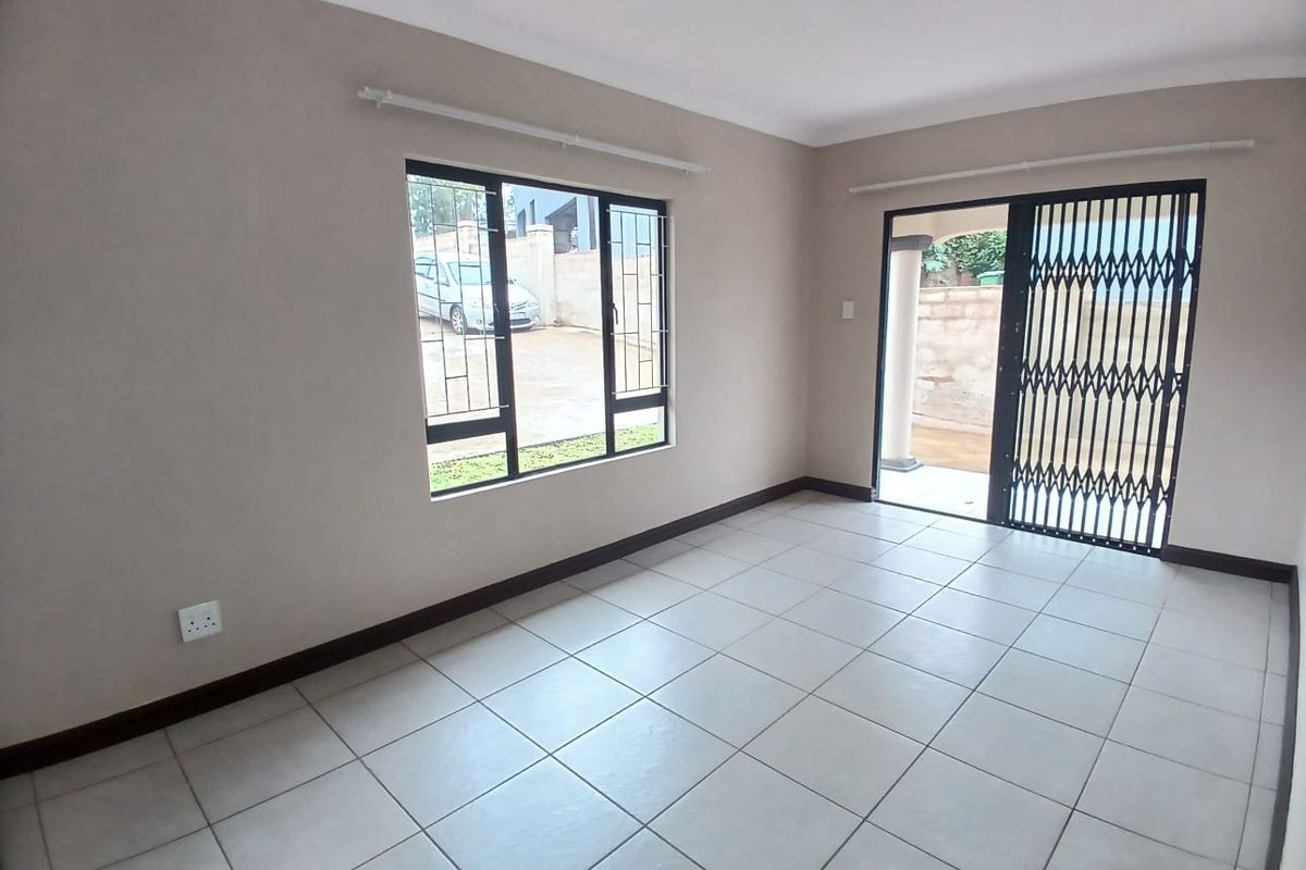 A bright, empty living room featuring neutral tiled flooring, security bars on the windows, and a sliding door leading to an outdoor area.