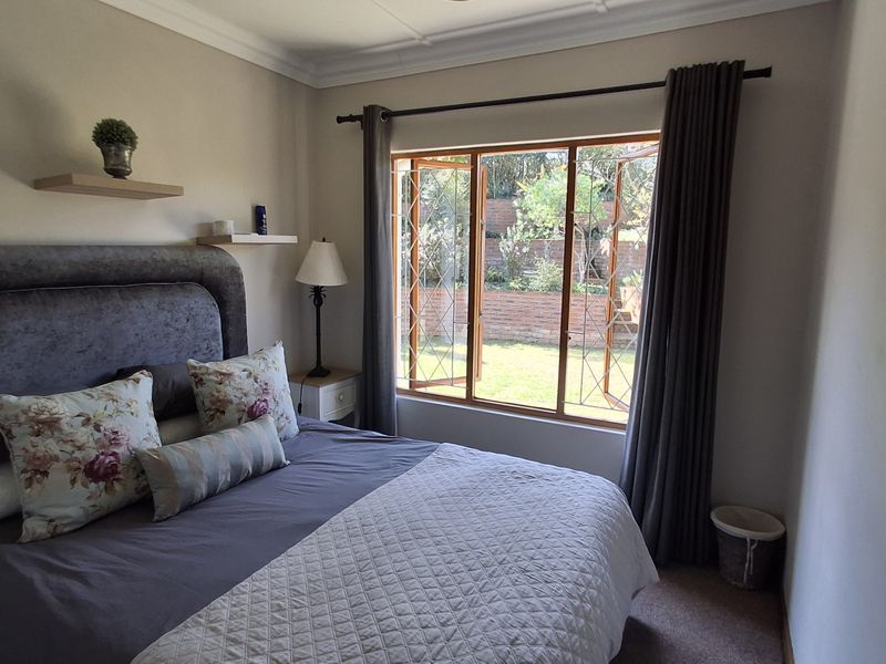 A well-maintained bedroom featuring timber-framed windows with security bars, floating wall shelves, and decorative cornice detailing, overlooking a private garden area.