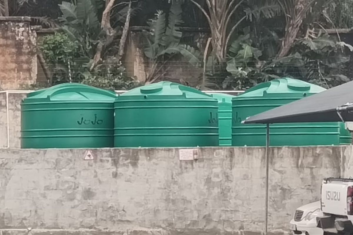 Exterior view showing a row of green water storage tanks positioned behind a weathered concrete retaining wall.
