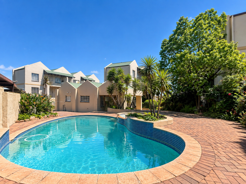 A well-maintained communal swimming pool area featuring paved decking, landscaped garden beds, and views of the surrounding apartment complex buildings.