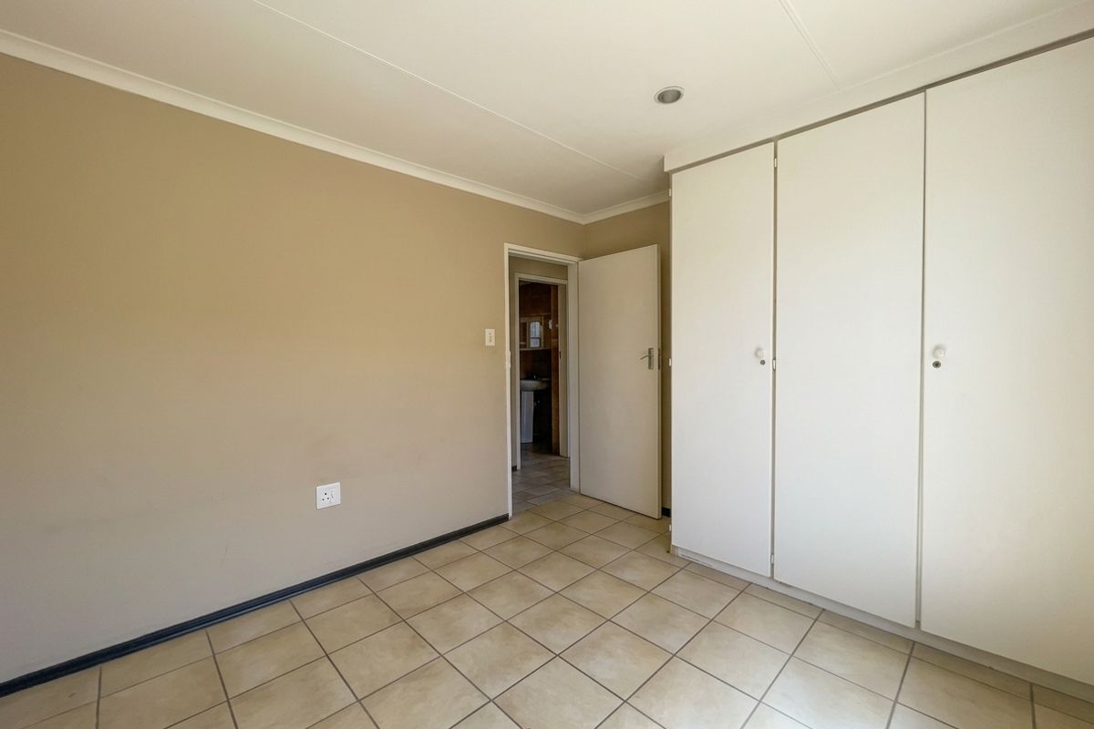 A clean, empty bedroom featuring neutral wall tones, tiled flooring, and built-in storage cupboards, with a view into the adjacent bathroom.