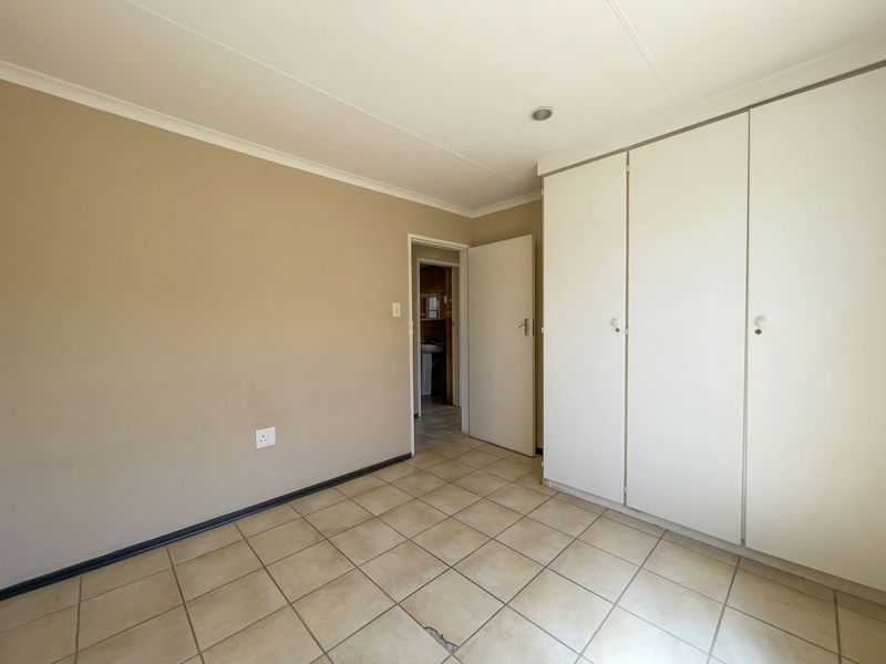 A clean, empty bedroom featuring neutral wall tones, tiled flooring, and built-in storage cupboards, with a view into the adjacent bathroom.