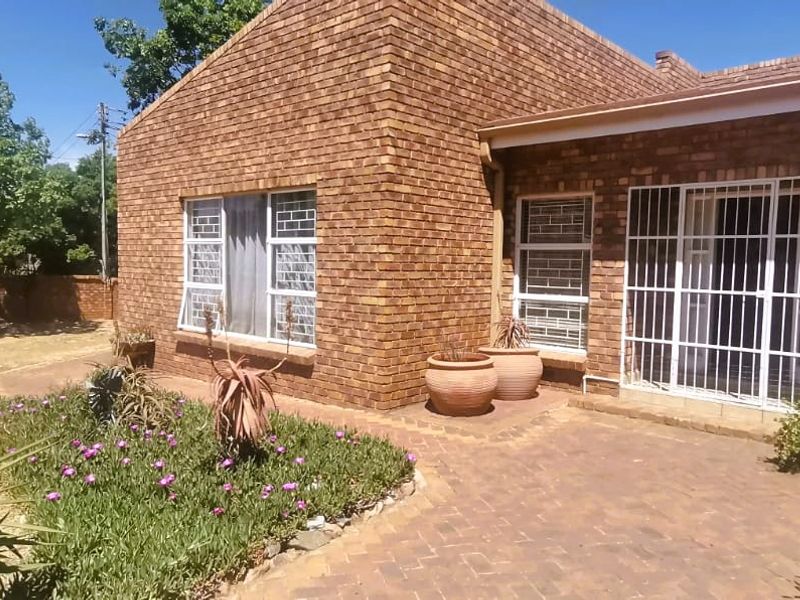 A well-maintained face brick rear exterior featuring a paved patio area and established garden beds with security bars on windows and doors.