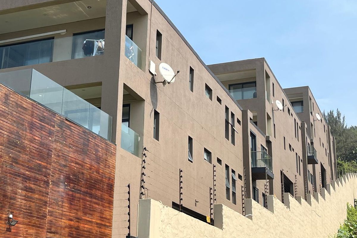 A modern multi-story apartment building exterior featuring glass balconies, timber cladding accents, and secure perimeter electric fencing.