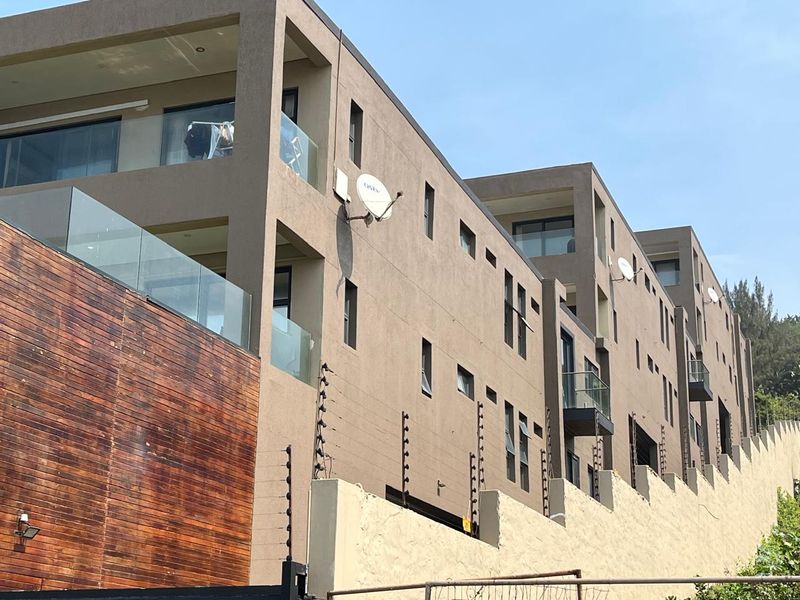 A modern multi-story apartment building exterior featuring glass balconies, timber cladding accents, and secure perimeter electric fencing.