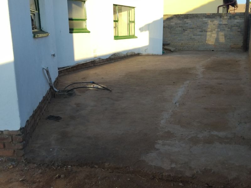 A rear exterior area featuring a concrete patio surface, brick boundary walls, and steel-framed windows, currently requiring maintenance and surface repairs.