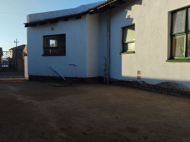 Rear exterior view of the property showing white painted walls, metal window frames, and a dirt yard area requiring maintenance.