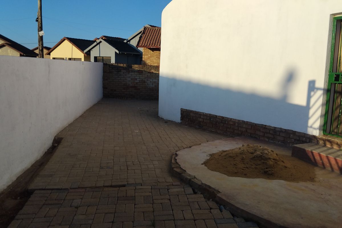A rear exterior area featuring paved surfaces, boundary walls, and a security gate, currently showing signs of unfinished landscaping and maintenance.