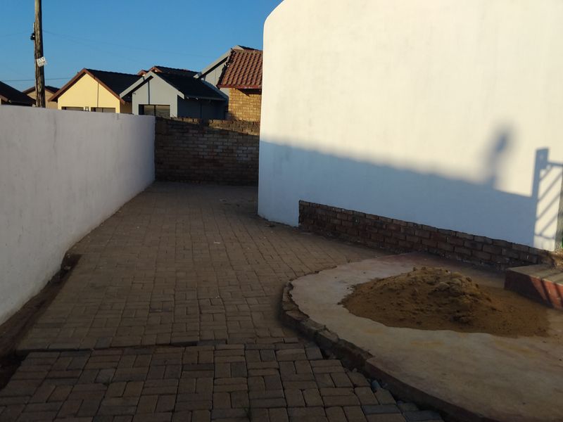 A rear exterior area featuring paved surfaces, boundary walls, and a security gate, currently showing signs of unfinished landscaping and maintenance.