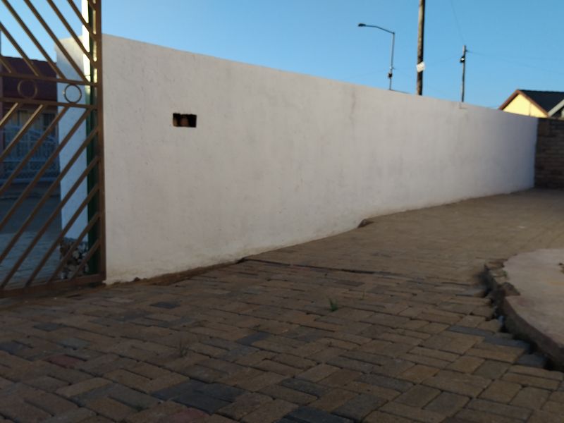 A view of the property entrance featuring a paved driveway, a metal gate, and a white boundary wall with a small section of missing brickwork.