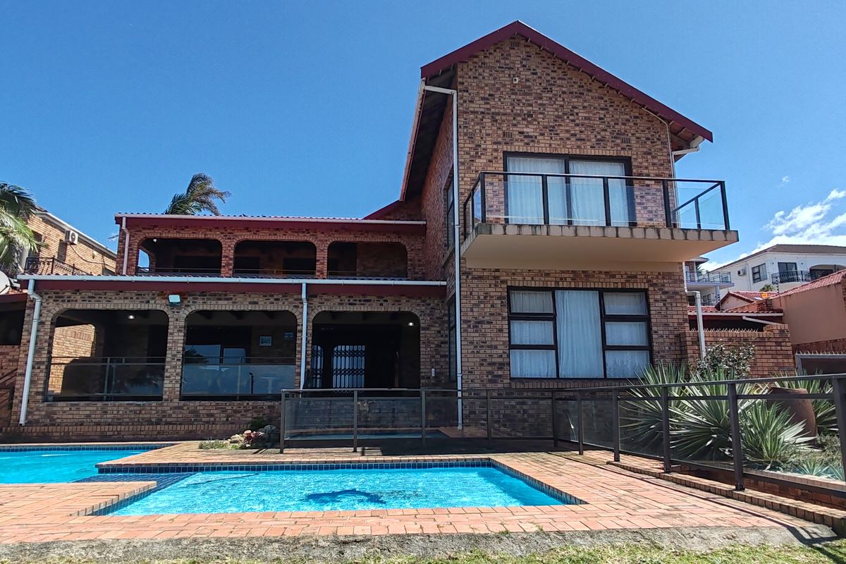 Rear view of a multi-story brick house featuring a swimming pool, covered patio, and an upper-level balcony with glass balustrades.