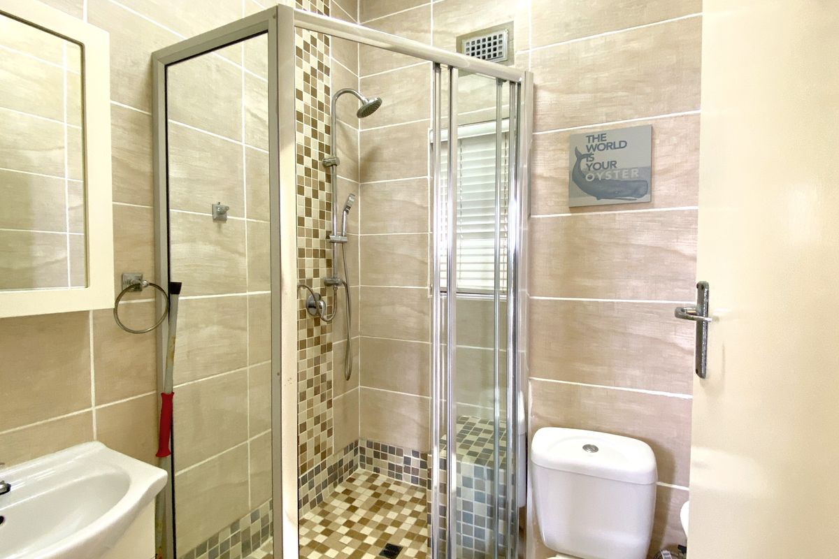 A compact bathroom featuring a glass-enclosed shower, pedestal basin, and toilet with neutral wall tiling and decorative mosaic accents.
