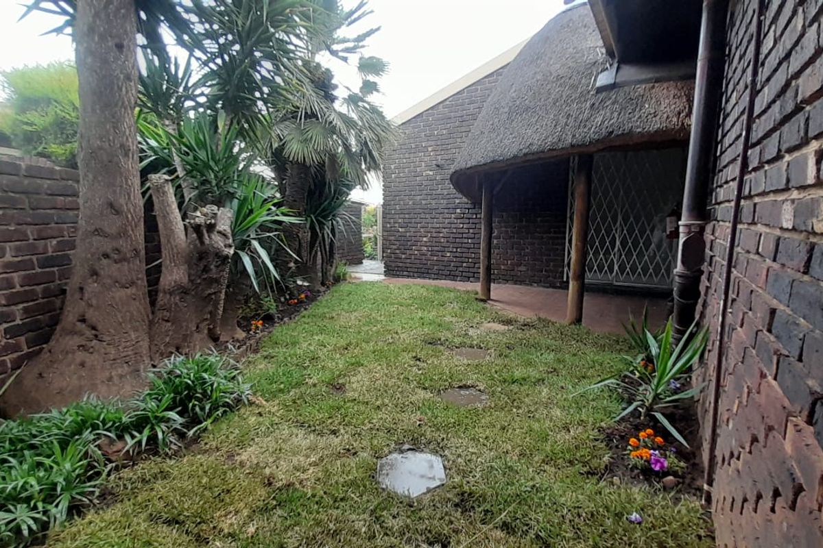 A rear garden area featuring a thatched roof overhang, brick exterior walls, and a small paved patio space with established trees.