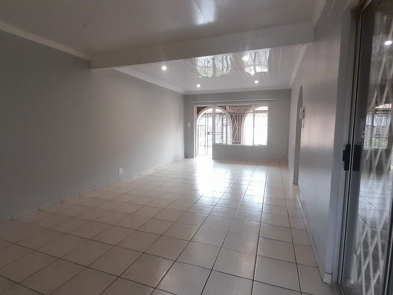 A spacious, empty living area featuring neutral tiled flooring, recessed lighting, and a sliding glass door with security gate leading to the exterior.