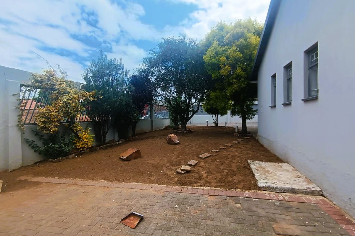 A rear exterior view featuring a paved area, boundary walls with security fencing, and an open garden space with mature trees.