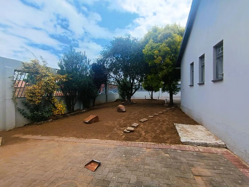 A rear exterior view featuring a paved area, boundary walls with security fencing, and an open garden space with mature trees.