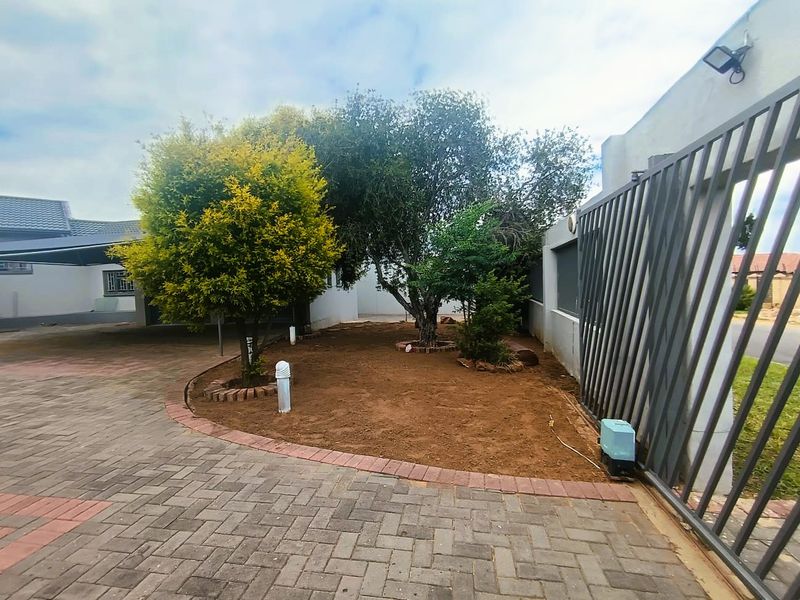 Front exterior view featuring a paved driveway, sliding gate with motor, and established trees within a landscaped garden area.