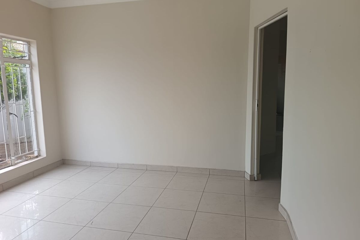 A bright, empty bedroom featuring neutral tiled flooring, clean walls, and a window fitted with security bars.
