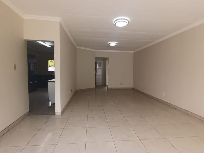 A spacious, empty living area featuring neutral tiled flooring, coved cornicing, and flush mount ceiling lighting, with an adjacent view into the kitchen area.