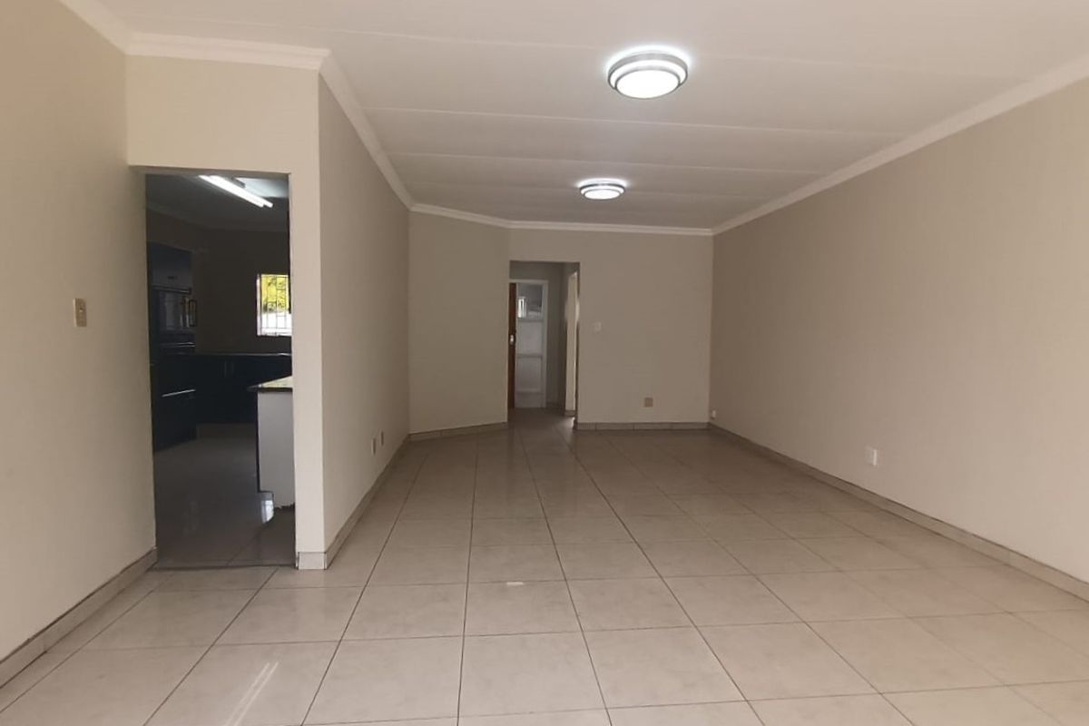 A spacious, empty living area featuring neutral tiled flooring, coved cornicing, and flush mount ceiling lighting, with an adjacent view into the kitchen area.