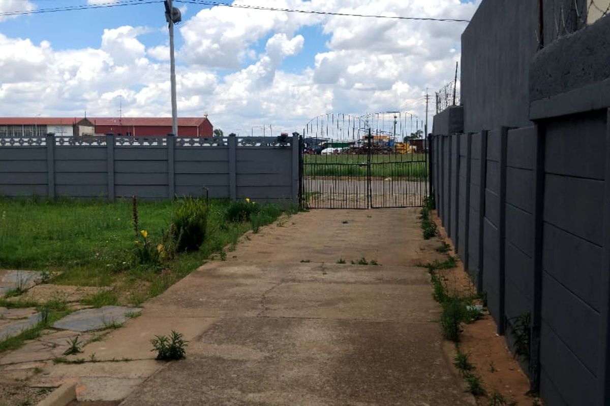 A rear exterior view featuring a concrete driveway, enclosed by precast concrete boundary walls and a metal gate.
