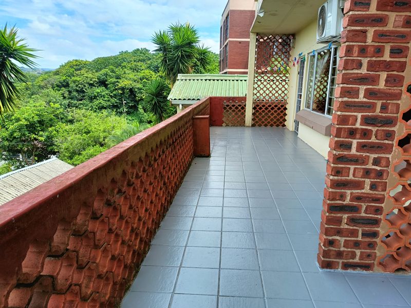 A spacious covered balcony featuring tiled flooring, decorative brick balustrades, and a scenic view of the surrounding greenery.