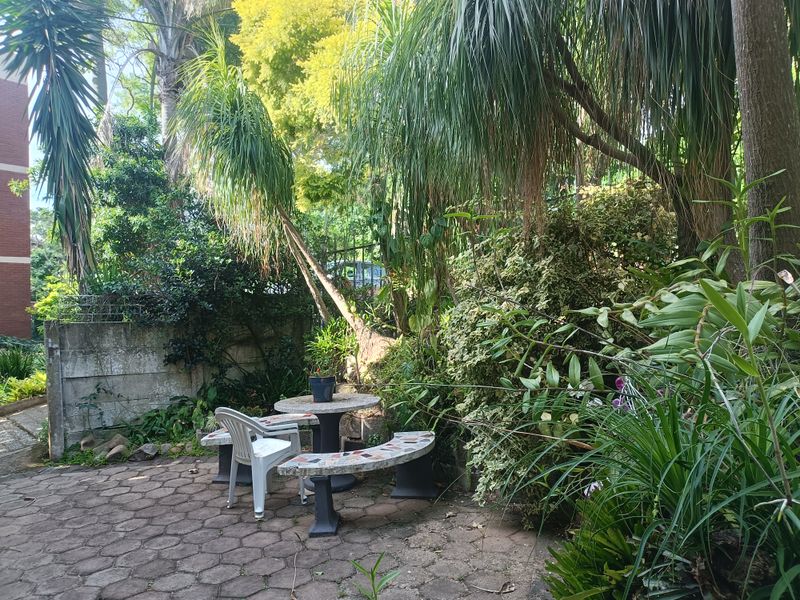 A private garden area featuring mature trees, lush greenery, and a paved patio space enclosed by a boundary wall.