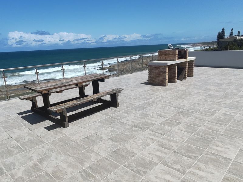 Spacious outdoor balcony featuring tiled flooring, a built-in brick braai, and a stainless steel balustrade with panoramic ocean views.
