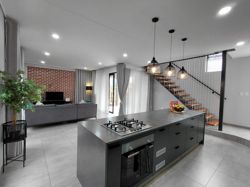 Modern open-plan living area featuring a sleek kitchen island with gas hob, integrated oven, and a striking staircase with contemporary pendant lighting.