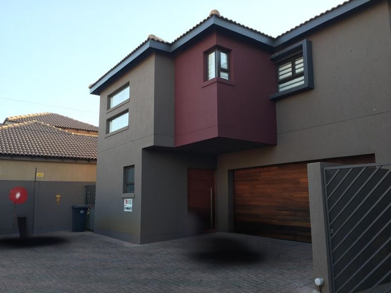 Modern double-storey townhouse exterior featuring a paved driveway, wooden garage door, and contemporary dark-toned facade with contrasting red accent detailing.