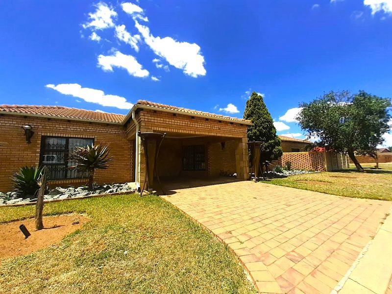 Front view of a single-story brick house featuring a paved driveway, covered entrance, and established garden area.