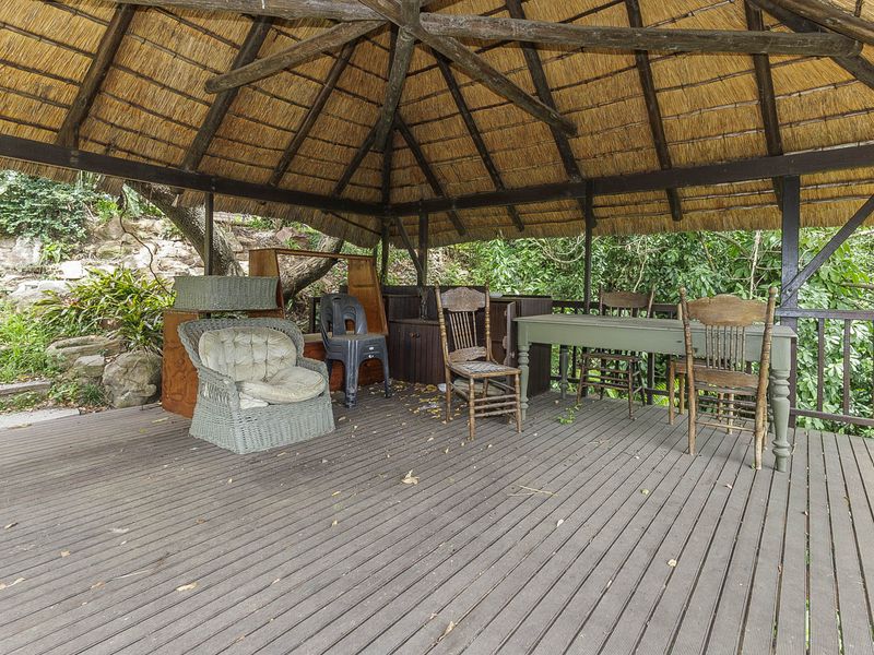 A rustic outdoor patio area featuring a traditional thatched roof, exposed timber rafters, and timber decking, set within a natural garden environment.
