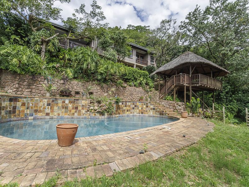 Outdoor pool area featuring a stone retaining wall, brick paving, and a raised thatched gazebo set against a lush, elevated garden backdrop.