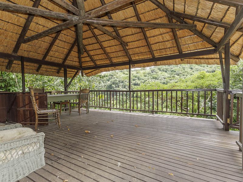 A spacious covered patio featuring a traditional thatched roof, timber decking, and a wooden balustrade, offering elevated views of the surrounding natural landscape.