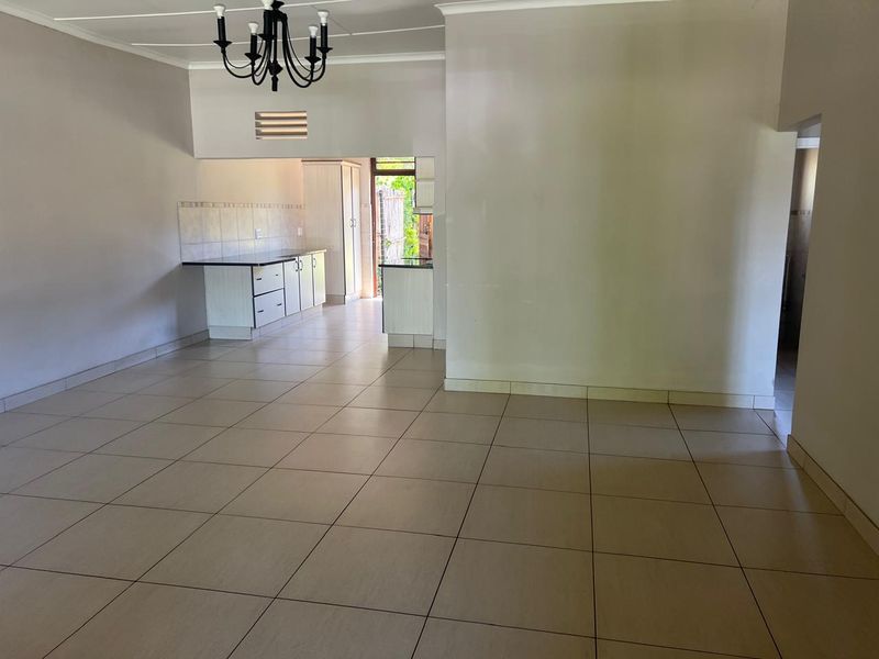 Spacious open-plan living area featuring neutral tiled flooring, a coved ceiling with a chandelier, and an adjoining kitchen space with built-in cabinetry.