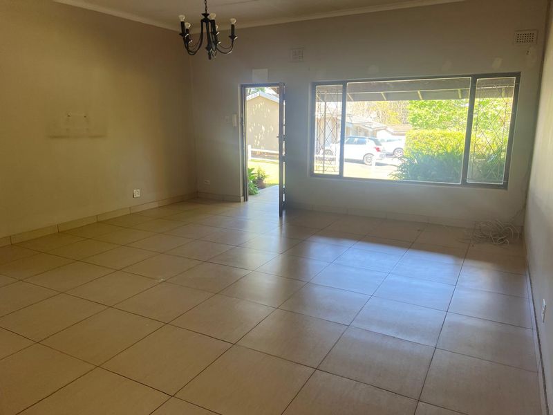 A spacious, empty living room featuring neutral tiled flooring, a large window with security bars, and direct access to the exterior.