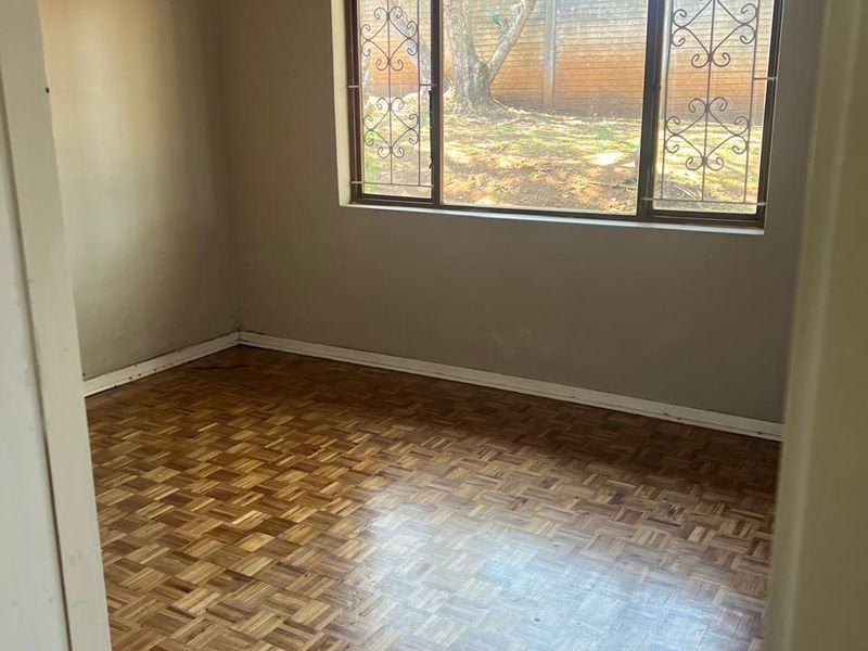 An empty bedroom featuring classic parquet flooring, large windows with security bars, and neutral wall tones, offering a blank canvas for personalization.