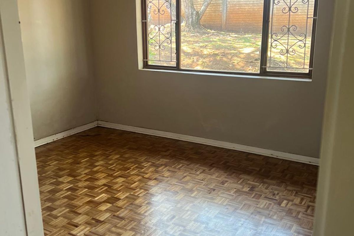 An empty bedroom featuring classic parquet flooring, large windows with security bars, and neutral wall tones, offering a blank canvas for personalization.