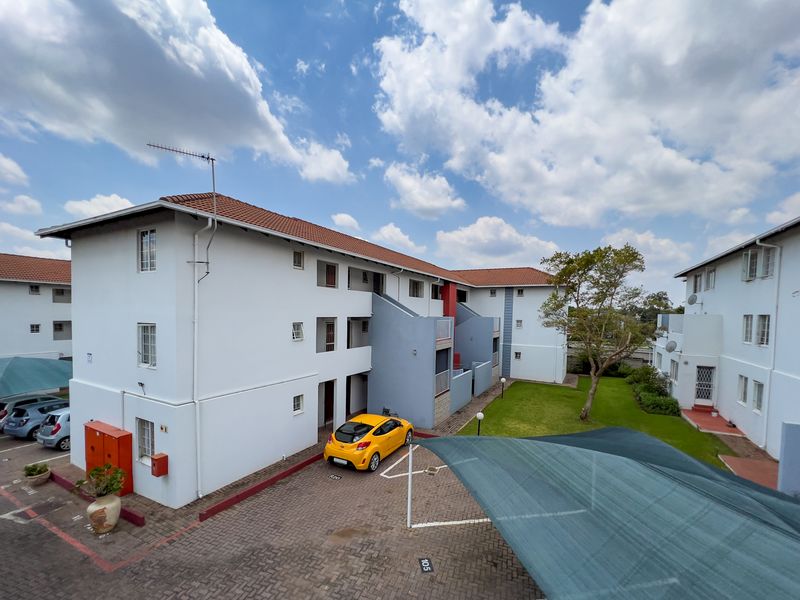 Exterior view of an apartment complex featuring a paved parking area with shade cloth carports, communal garden space, and multi-story residential building blocks.