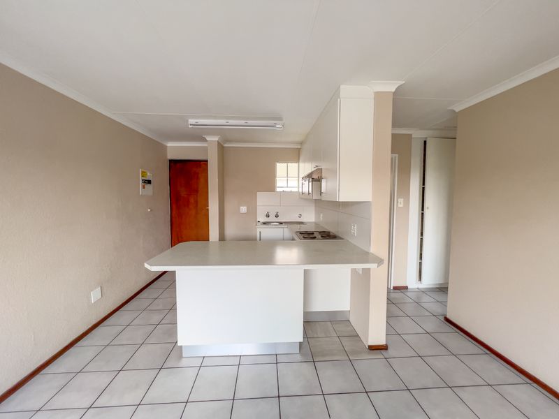 A functional, open-plan kitchen featuring white built-in cabinetry, a laminate breakfast bar, electric hob, and tiled flooring throughout.