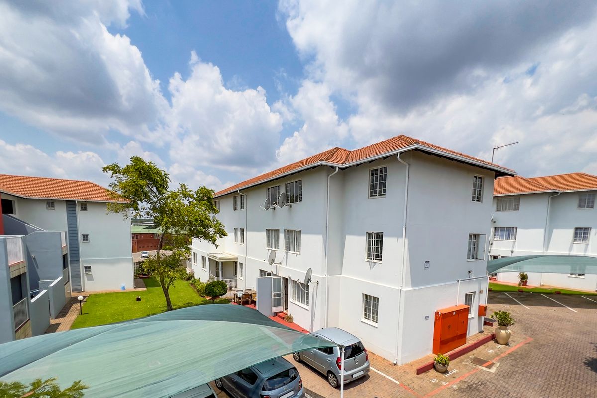 Exterior view of a multi-story apartment complex featuring a tiled roof, paved parking area with shade cloth carports, and a communal garden space.